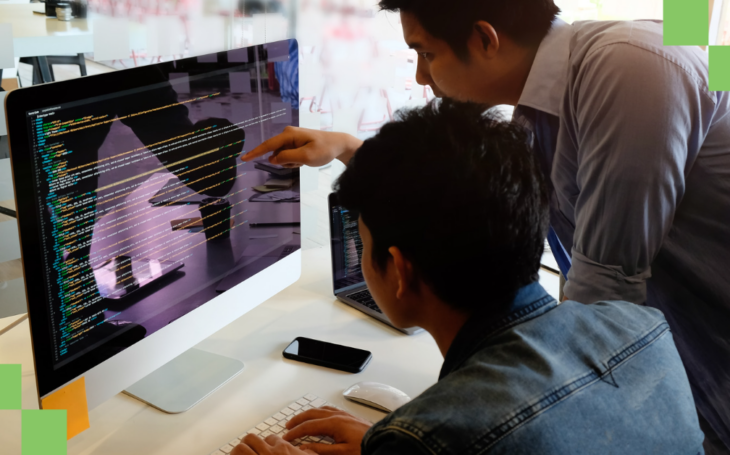 two-male-coworkers-leaning-over-desk-looking-at-lines-of-code-on-a-computer-monitor-discussing-task-automation